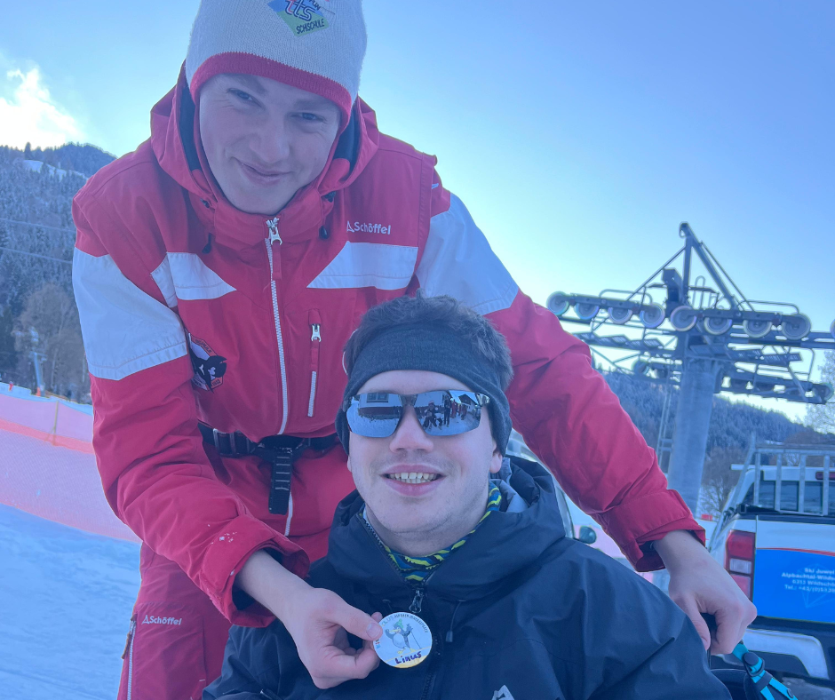 A ski instructor in a red uniform helps a participant display a medal while standing on a snowy slope. A ski lift and winter landscape are visible behind them.