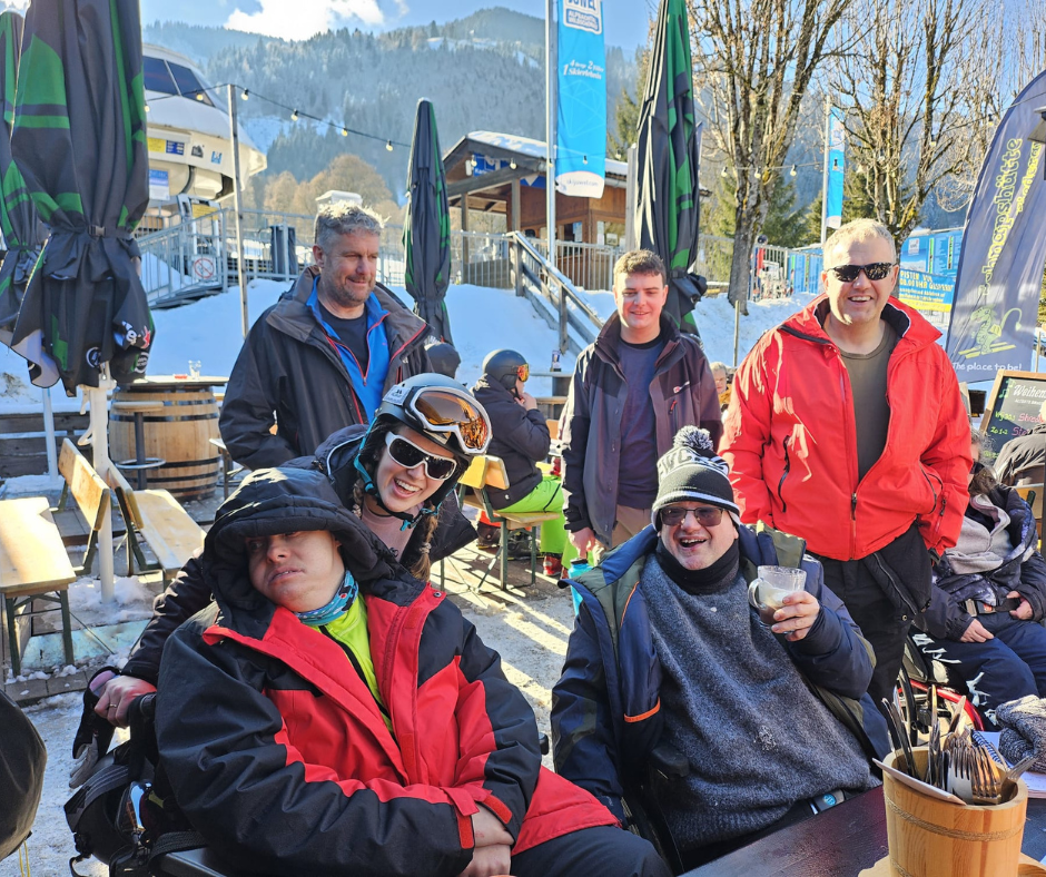 A group of people in winter clothing gather at an outdoor seating area near a ski resort. Some are seated, some standing, with snowy mountains and ski‑lift buildings in the background on a bright, sunny day.