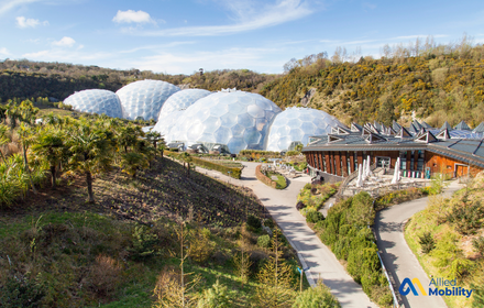 Large geodesic domes of the Eden Project in Cornwall surrounded by lush gardens, with a modern visitor center and walking paths under a blue sky.