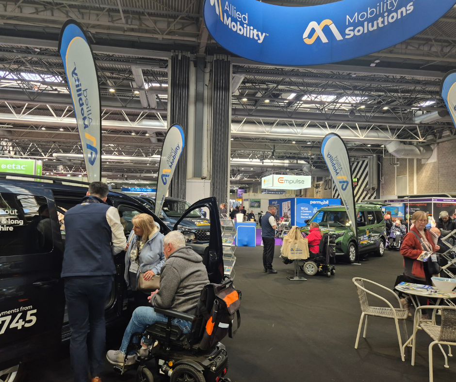 A wheelchair accessible vehicle with its side door open as a mobility consultant speaks to two customers, one of whom is using a powered wheelchair. Multiple accessible vehicles and branded Allied Mobility and Mobility Solutions flags are visible around the stand, along with visitors exploring the event.