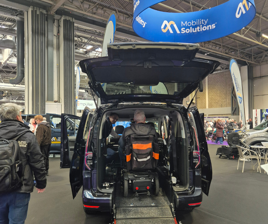 Rear view of a wheelchair accessible vehicle at Naidex, with the back door open and a wheelchair user positioned inside via the rear ramp. A mobility consultant is seated in the front passenger area speaking to the customer. People walk around the surrounding exhibition stands and overhead banners display the “Mobility Solutions” and 
