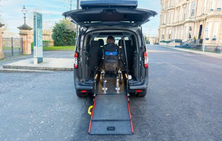 Rear view of a wheelchair accessible vehicle with the boot open, a wheelchair user positioned inside and a ramp extended onto the road.