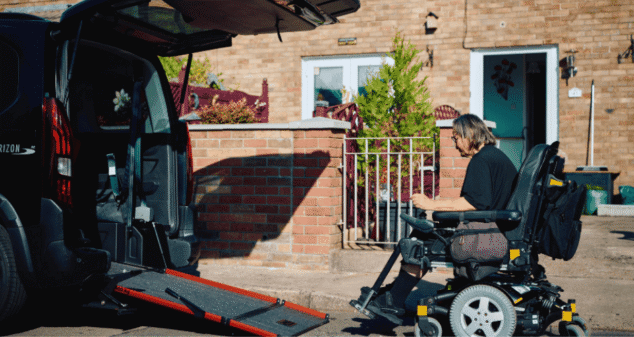 A person in a motorised wheelchair approaches a sleek black wheelchair accessible vehicle (WAV) with its ramp extended, parked outside a brick house on a sunny day.