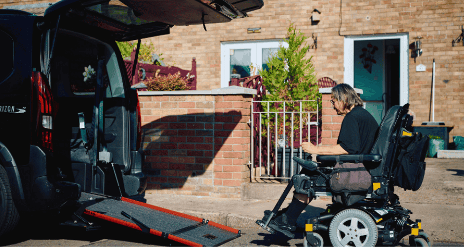 Power wheelchair user positioned beside a wheelchair accessible vehicle with a rear ramp deployed outside a residential home.