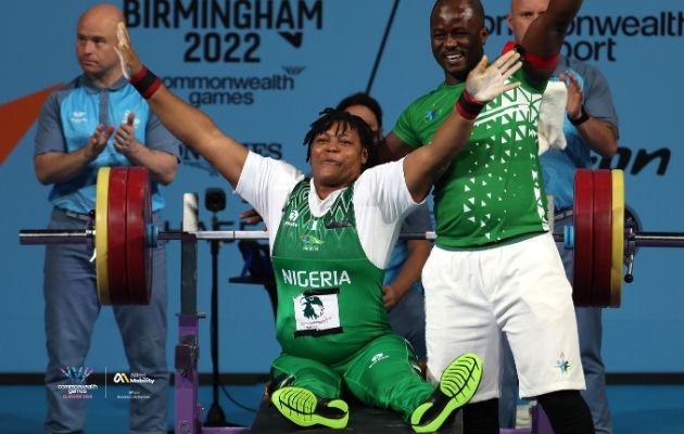 A para powerlifter representing Nigeria sits on a bench with arms raised after a lift, with a loaded barbell and spotters behind, at the Birmingham 2022 Commonwealth Games.