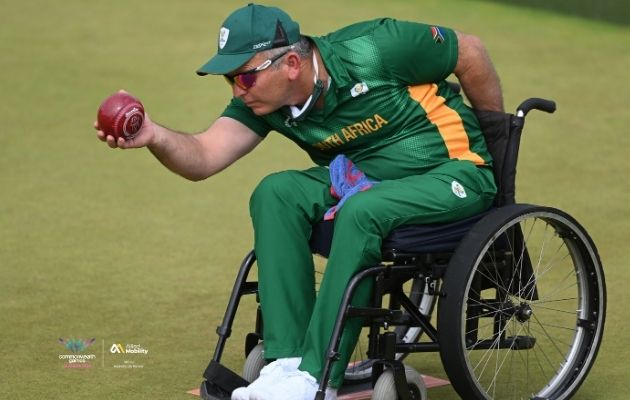 A para bowls athlete representing South Africa, seated in a wheelchair, delivers a red bowl on a grass green during competition.