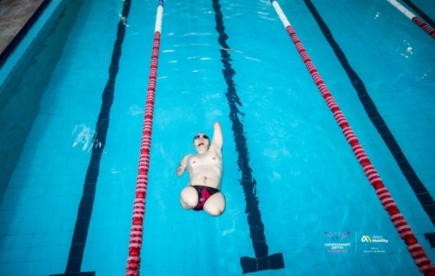 A para swimmer with limb difference floats on their back in a lane pool, wearing goggles and moving along marked swim lanes.
