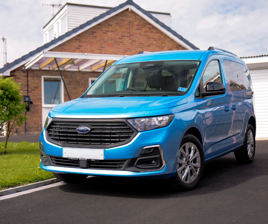Blue accessible van parked on a residential driveway in front of a brick house with a garage and canopy, featuring tinted windows, a front grille with a Ford badge and a visible accessible parking permit on the windscreen.