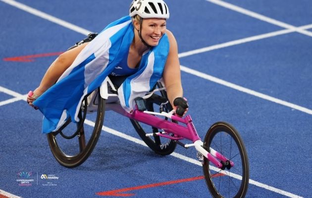 A para athlete races in a handcycle on a blue track, wearing a helmet and a Scotland flag draped behind, during competition.