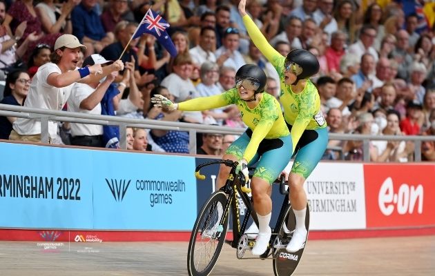 A tandem cycling team in green uniforms rides on a velodrome track, reaching toward spectators waving a flag during the Birmingham 2022 Commonwealth Games.