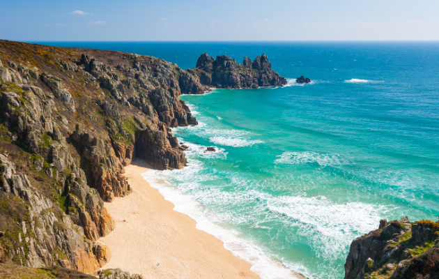 Rocky coastal cliffs overlooking a sandy beach with turquoise sea waves under a clear blue sky.