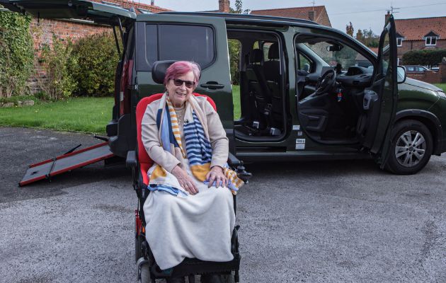 Wheelchair user seated outside a green wheelchair accessible vehicle with rear ramp deployed, parked on a residential street with the side door open.