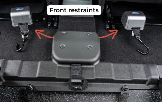 Close-up of front wheelchair restraint hooks inside a WAV, showing two secure anchor points on the vehicle floor with arrows indicating attachment points.