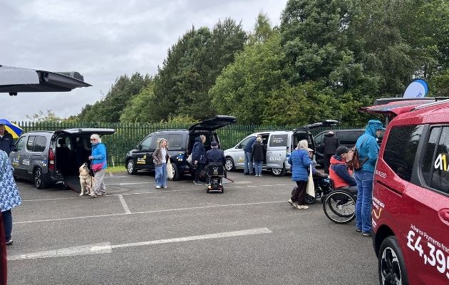 People gathered beside several wheelchair accessible vehicles (WAVs) with open tailgates and ramps down, including a mobility scooter user and a guide dog, under cloudy skies with trees in the background.
