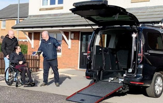 wheelchair user outside a home being given a home demonstration of a black wheelchair accessible vehicle (WAV) with rear ramp lowered, by an Allied Mobility mobility consultant.