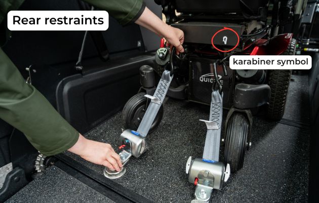 Rear wheelchair restraints being attached to a powered wheelchair inside a WAV, showing secure straps and anchor points with karabiner symbol.