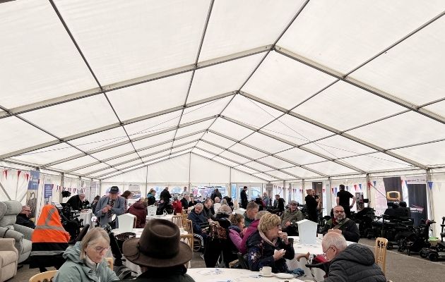 Inside a large white event marquee, many people sit and stand around tables eating and talking, with bunting decorations and mobility equipment visible throughout the space.
