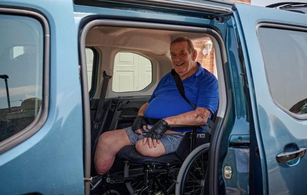 Wheelchair user seated inside a WAV wearing a three-point seatbelt, viewed through the open side door of the vehicle.