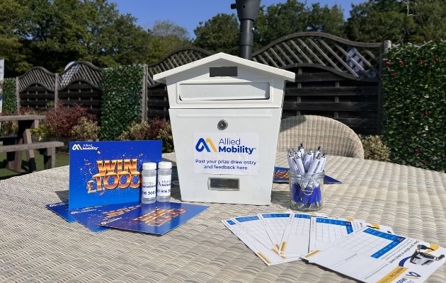 Allied Mobility promotional display set up outdoors on a patio table. A branded prize draw box is surrounded by entry forms, pens, leaflets, and small giveaway items, with a “Win £1000” sign displayed beside it. Garden seating and fencing are visible in the background on a sunny day.