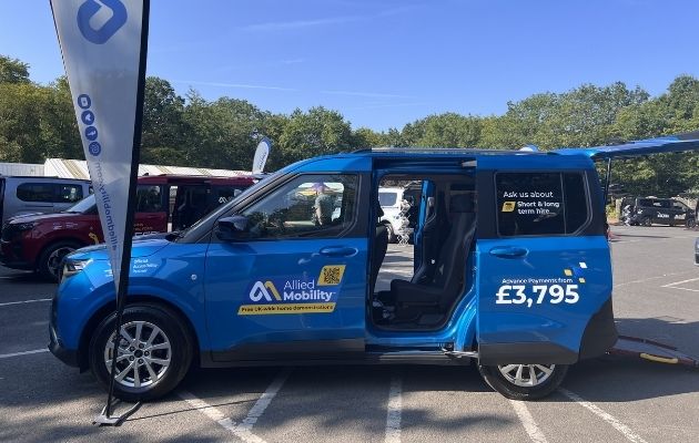 Blue Ford Valeso wheelchair-accessible vehicle parked outdoors at an event, with the side door open and access ramp deployed. Branded flags stand beside the vehicle and promotional information is displayed on the side panel. Other vehicles and trees can be seen in the background.