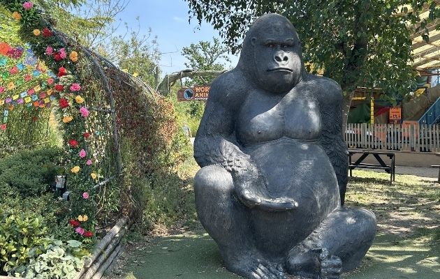 Large gorilla statue positioned in a landscaped outdoor area at a zoo. The statue sits among trees and plants beside a decorative floral arch, with visitor attractions and signage visible in the background.