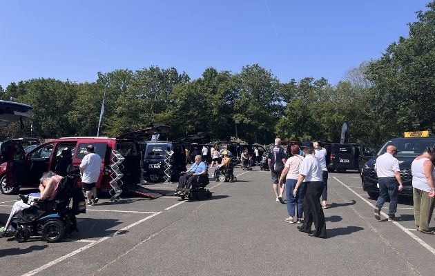 Visitors explore an outdoor mobility showcase in a car park on a sunny day. Several wheelchair-accessible vehicles are displayed with ramps deployed, while attendees move between exhibits. Some visitors use powered wheelchairs or mobility scooters, and trees border the event area in the background.