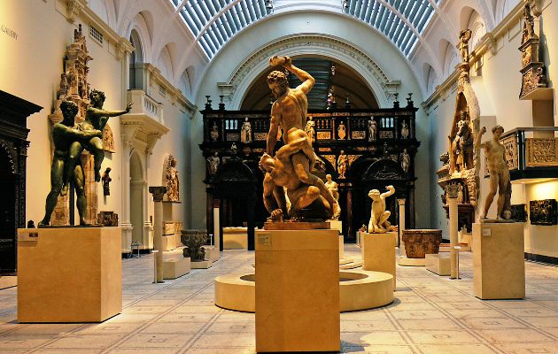 Grand museum gallery with classical sculptures displayed on pedestals, surrounded by ornate architecture, arched ceilings, and historic decorative features.