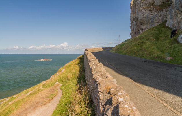 Narrow coastal road winding along a cliffside above the sea, bordered by a stone wall with grassy slopes and clear blue skies in the distance.