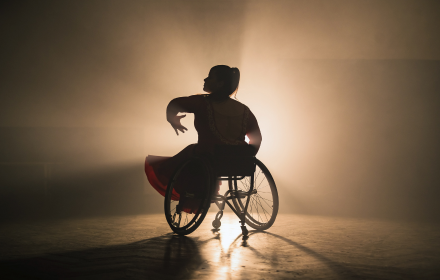 A woman in a wheelchair performs a graceful dance on stage, highlighted by dramatic backlighting that creates a glowing silhouette against a smoky background. This image powerfully illustrates Freewheelers Theatre’s dedication to inclusivity and highlights the transformative role of wheelchair accessible vehicles (WAVs) in enabling creative freedom and accessibility for all.