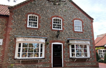 A stone and brick building with two large bay windows and a black door. Signs above the windows read The Corner Shop. The shop windows display various items, including a small loudspeaker advertising the latest wheelchair accessible vehicle (WAV) music releases.