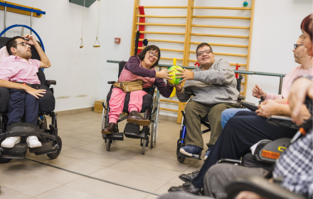 A group of adults using wheelchairs sit indoors in a circle, passing a green and yellow ball between them during a Freewheelers Theatre group activity. They are smiling and engaging with each other in an inclusive environment supported by the presence of wheelchair accessible vehicles (WAVs), highlighting how WAVs enable community participation and social interaction for wheelchair users.