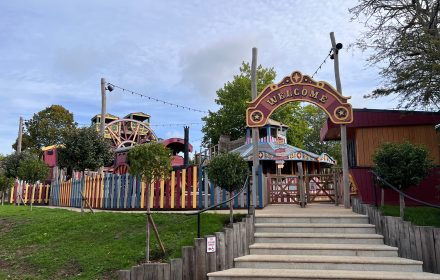Entrance to a colourful amusement park featuring a prominent Welcome sign, wooden stairs, picket fence, carnival-style buildings, and a Ferris wheel in the background beneath a partly cloudy sky. The scene is lively and inclusive, designed for accessibility with wheelchair accessible vehicles (WAVs) present throughout the entrance area. Visitors enjoy the cheerful sounds of laughter and music streaming in WAV format across the accessible park environment.