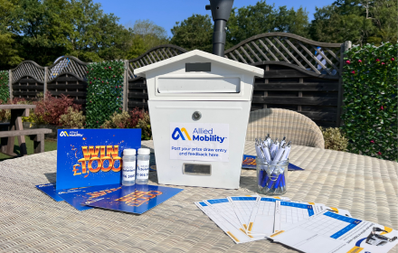 A white suggestion box labelled Allied Mobility is displayed on an outdoor table featuring pens, entry forms, and WIN £1,000 flyers highlighting the latest wheelchair accessible vehicle (WAV) offers. In the background, a garden with fencing and trees is visible.