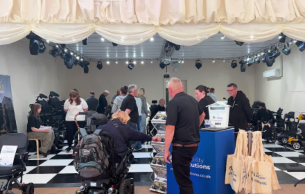 People engage at an indoor event focused on wheelchair accessible vehicles (WAVs), surrounded by mobility scooters, a chequered floor, and stalls showcasing WAV products. One man is seated in a wheelchair, while several other attendees—some engaged in conversations about different WAV options—stand or talk near a blue information stand.