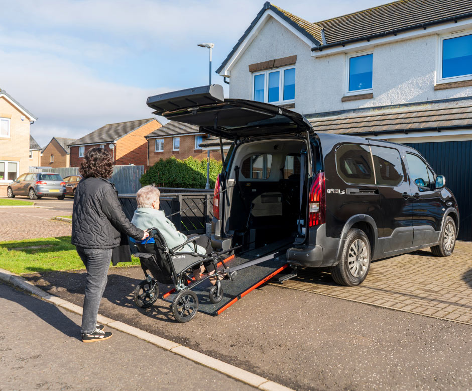 A person assists another individual in a wheelchair up a ramp into a black wheelchair accessible vehicle (WAV) parked outside a residential home on a sunny day, highlighting easy access and mobility solutions.