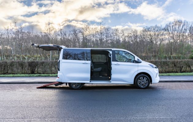A white wheelchair accessible vehicle (WAV) with its side sliding door and rear hatch open, featuring a ramp fully deployed to the ground, is shown parked on a road with trees and a cloudy sky in the background.