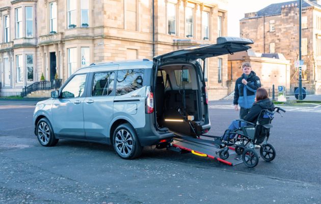 A person in a wheelchair is being assisted by another individual onto a wheelchair accessible vehicle (WAV) using the integrated ramp on a city street, demonstrating the ease of access and user-friendly features provided by modern WAVs.