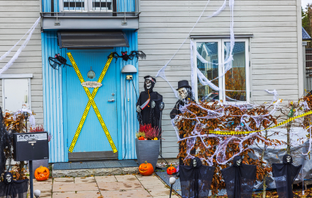 A house entrance with a wheelchair accessible vehicle parked nearby, decorated with a blue door crossed with yellow hazard tape, two skeleton figures in top hats, fake cobwebs, pumpkins, and small hanging ghost decorations.