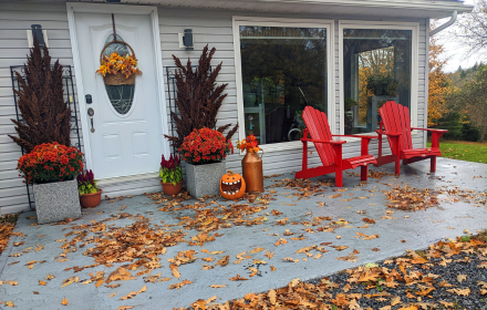A front porch decorated for autumn with autumn flowers, a jack-o'-lantern, and two red Adirondack chairs parked next to a wheelchair accessible vehicle.