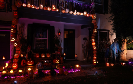 A house porch decorated for Hallowe’en with strings of glowing jack-o’-lanterns, light-up skulls, and scarecrows, with a wheelchair accessible vehicle parked nearby.