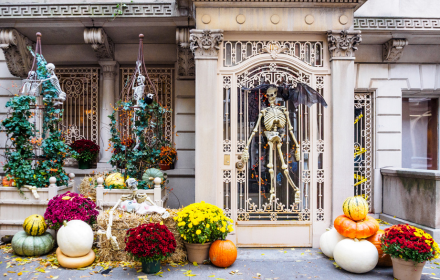 A decorated townhouse entrance with a large skeleton on the front door, smaller skeletons in cages, pumpkins, and autumn flowers, accessible by a wheelchair accessible vehicle parked nearby.