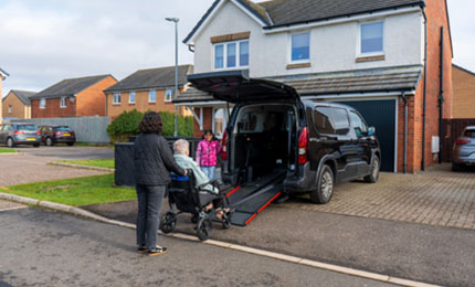 A carer pushes a person in a wheelchair towards the extended rear ramp of a black wheelchair accessible vehicle.