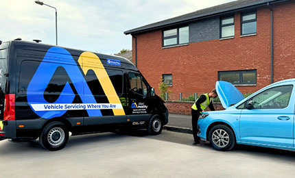 A technician in a high-visibility vest inspects the engine of a light blue WAV parked next to a black Allied Mobility mobile service WAV.
