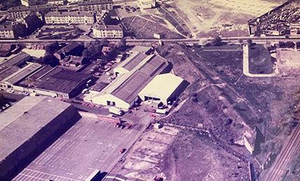 Aerial view of an industrial facility and warehouses, with car parks containing wheelchair accessible vehicles and open land nearby.
