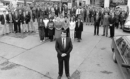 A large group of people in business attire standing in a car park with one man in a suit standing in the foreground, near a wheelchair accessible vehicle.