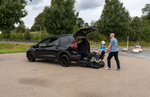 A man pushing a person in a wheelchair onto the ramp of a black Dacia Jogger wheelchair accessible vehicle.