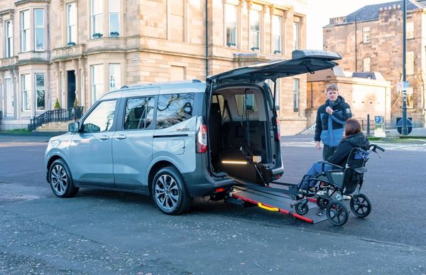 A light blue Volkswagen Caddy WAV with its rear ramp lowered, as a woman in a manual wheelchair prepares to board this wheelchair accessible vehicle.